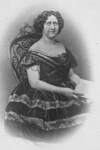 A woman in a 19th-century dress sits on an ornate chair, holding a paper.